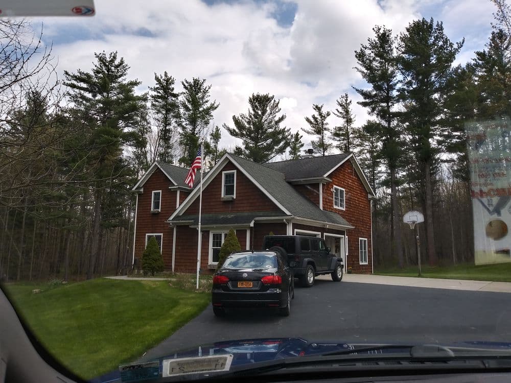 Brown house with American flag, surrounded by trees and parked cars in the driveway.