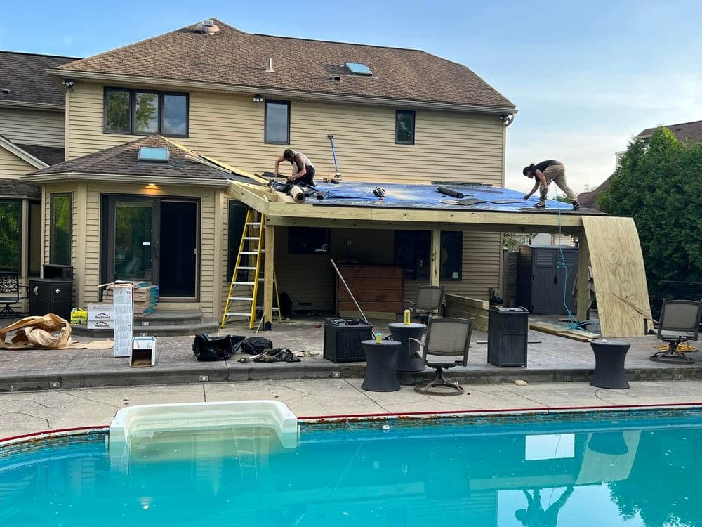 Roof installation in progress on a home patio with a pool in the foreground. Workers and tools visible.