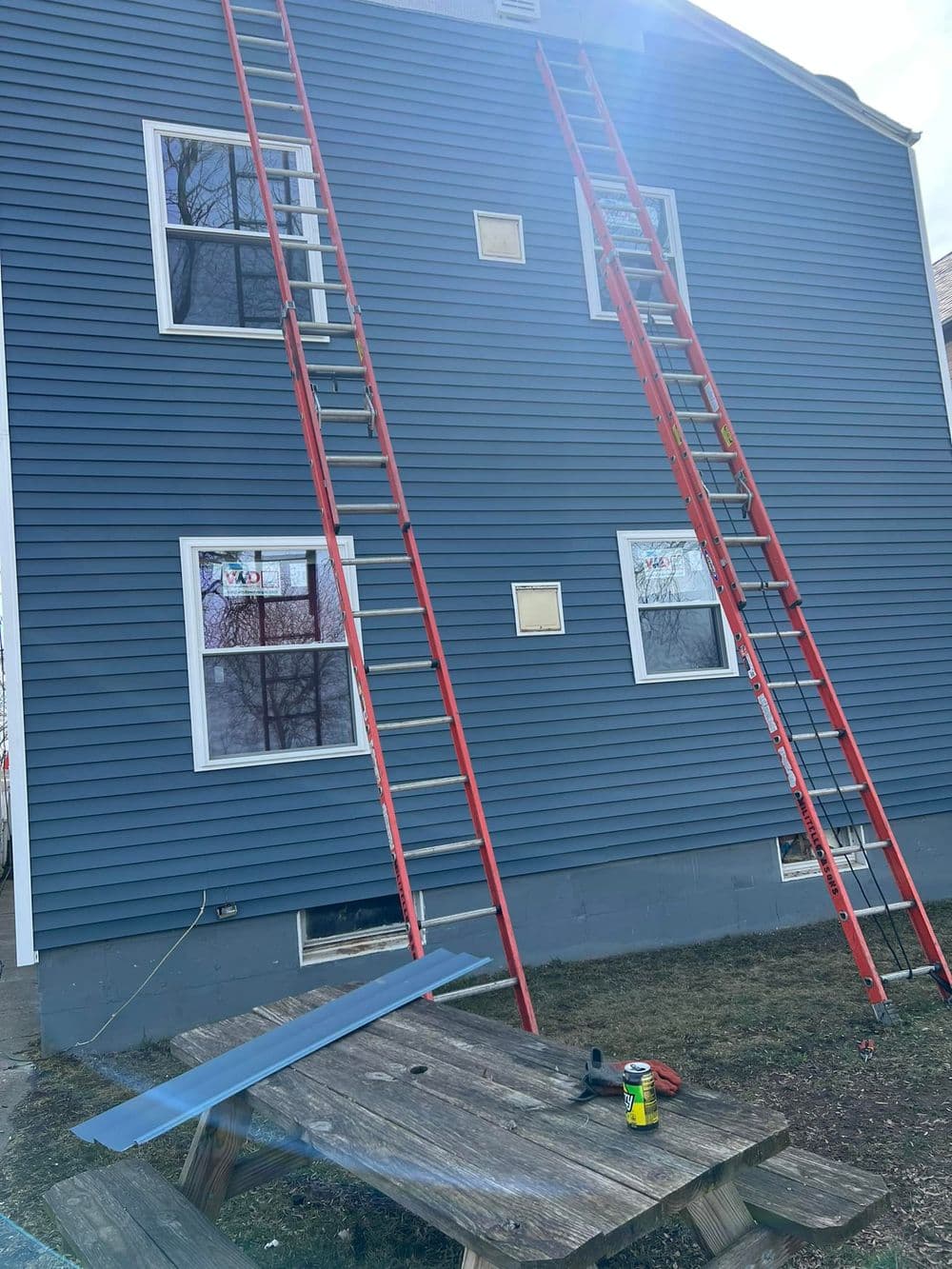 Blue house exterior with red ladders leaning against it, windows being replaced. Tools nearby.