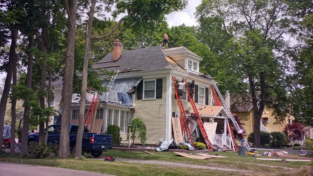 Workers repairing a house roof with ladders and tools surrounded by trees and parked truck.